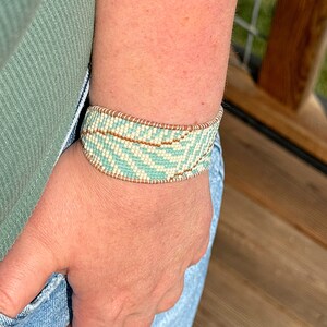 May include: A light blue and white beaded bracelet with a brown accent. The bracelet is worn on a person's wrist.