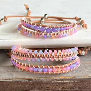 Op de afbeelding: Twee wikkelarmbanden met pastelkleurige kralen. De armbanden hebben een lichtbruine leren band met rijen roze, paarse en oranje kralen. De armbanden hebben verstelbare sluitingen met zilverkleurige metalen accenten.