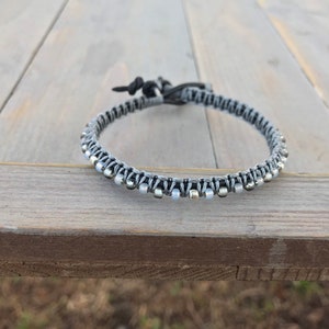 May include: A handmade bracelet featuring a woven design with gray and black cord. The bracelet is adorned with small, silver-colored beads. The adjustable closure is made of black cord. The bracelet is displayed on a wooden surface.