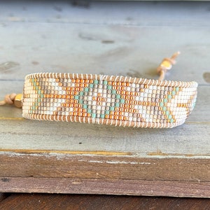 May include: A beaded bracelet with a geometric pattern in shades of white, beige, orange, and green. The bracelet is made with leather cord and has a gold clasp.