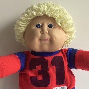 May include: A vintage Cabbage Patch Kid doll wearing a red jersey with the number 31, blue shorts, and white sneakers with blue stripes. The doll has blonde yarn hair and blue eyes. The doll's arms are outstretched.