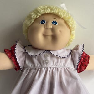 May include: A doll with light skin, blue eyes, and curly yellow yarn hair tied with a white ribbon. The doll wears a white short-sleeved dress with a collar, red polka-dot bloomers, and red shoes with small white flower accents.