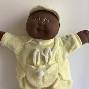 May include: A vintage Cabbage Patch Kid doll with dark skin, wearing a light yellow terrycloth outfit with a bunny-themed pocket and matching hat. The doll has white sneakers and gray socks. The doll's arms are outstretched.