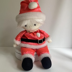 May include: A vintage Santa Claus doll with a red satin suit, a black belt, and black boots. The doll has white hair and a matching red hat with a white pom-pom. The hat and suit are trimmed with white faux fur.