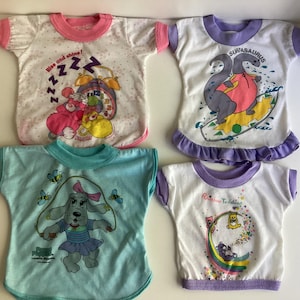 May include: Four different children's t-shirts with various cartoon characters and designs. The shirts are white with colorful graphics. The designs include a pink alarm clock, a dinosaur surfing, a dog jumping rope, and a rainbow with teddy bears.