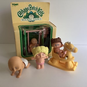 May include: A vintage Cabbage Patch Kids Rocking Babies toy set. The set includes a box with a baby doll on a swing, and three additional dolls, one crawling and one on a yellow rocking horse. The dolls have various hair colors and are wearing diapers.