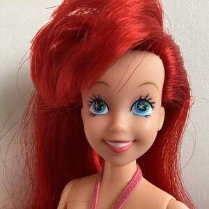 Peut inclure: Poup&eacute;e aux cheveux roux et aux yeux bleus. La poup&eacute;e porte un haut rose et affiche une expression souriante. Les yeux de la poup&eacute;e pr&eacute;sentent un motif en forme d'&eacute;toile. Les cheveux de la poup&eacute;e sont coiff&eacute;s de mani&egrave;re volumineuse.