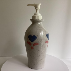 May include: A ceramic soap dispenser with a speckled light gray finish, featuring a white plastic pump mechanism. The body is decorated with hand-painted blue heart motifs and small pink curved accents arranged in a repeating pattern around the exterior.