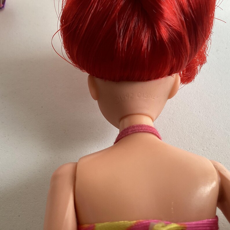 Peut inclure: Une poup&eacute;e avec des cheveux roux et un ras du cou rose. Le dos de la t&ecirc;te de la poup&eacute;e est visible, avec le texte "&copy;1992 Disney" imprim&eacute; au dos du cou. La poup&eacute;e porte une robe color&eacute;e.