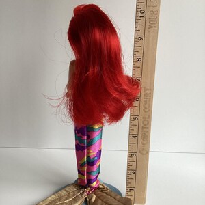 Peut inclure: Poup&eacute;e sir&egrave;ne avec de longs cheveux roux, portant une queue &agrave; motifs color&eacute;s et une nageoire caudale dor&eacute;e. La poup&eacute;e est pos&eacute;e sur une base bleue. Une r&egrave;gle en bois mesure jusqu'&agrave; 30 cm.