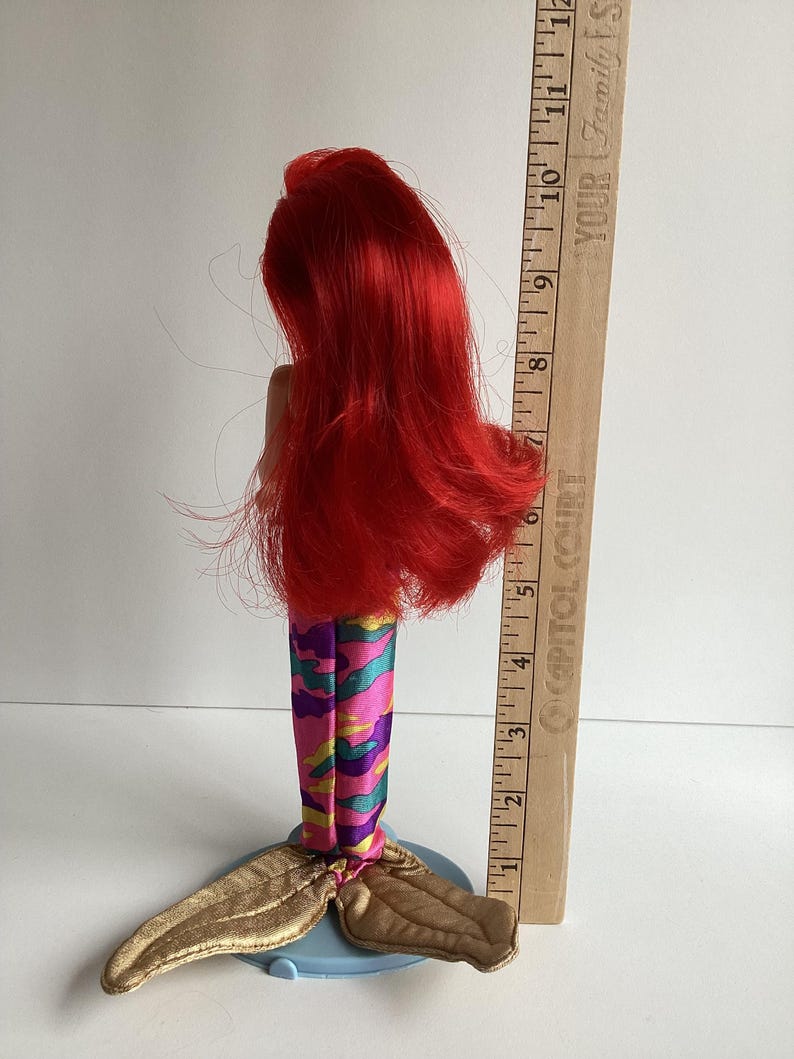 Peut inclure: Poup&eacute;e sir&egrave;ne avec de longs cheveux roux, portant une queue &agrave; motifs color&eacute;s et une nageoire caudale dor&eacute;e. La poup&eacute;e est pos&eacute;e sur une base bleue. Une r&egrave;gle en bois mesure jusqu'&agrave; 30 cm.