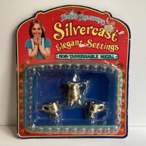 May include: A plastic package with a red background featuring the text "Terry's Treasures Silvercast Elegant Settings Non-Tarnishable Metal" and a picture of a young woman. The package contains a silver pitcher and two silver cups.