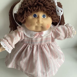May include: A vintage doll with brown yarn hair styled in pigtails with white ribbons. The doll has blue eyes and rosy cheeks. It wears a pink and white striped dress with a white collar and lace trim.
