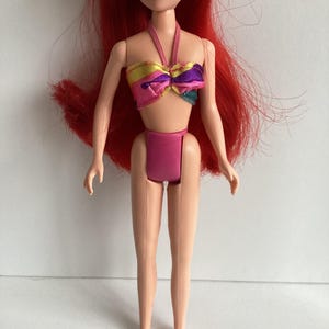 Peut inclure: Poup&eacute;e aux cheveux roux et aux yeux bleus, v&ecirc;tue d'un maillot de bain rose. Le haut de la poup&eacute;e est un style licou color&eacute; avec du jaune, du rose, du violet et du sarcelle. La poup&eacute;e est debout, les bras le long du corps.
