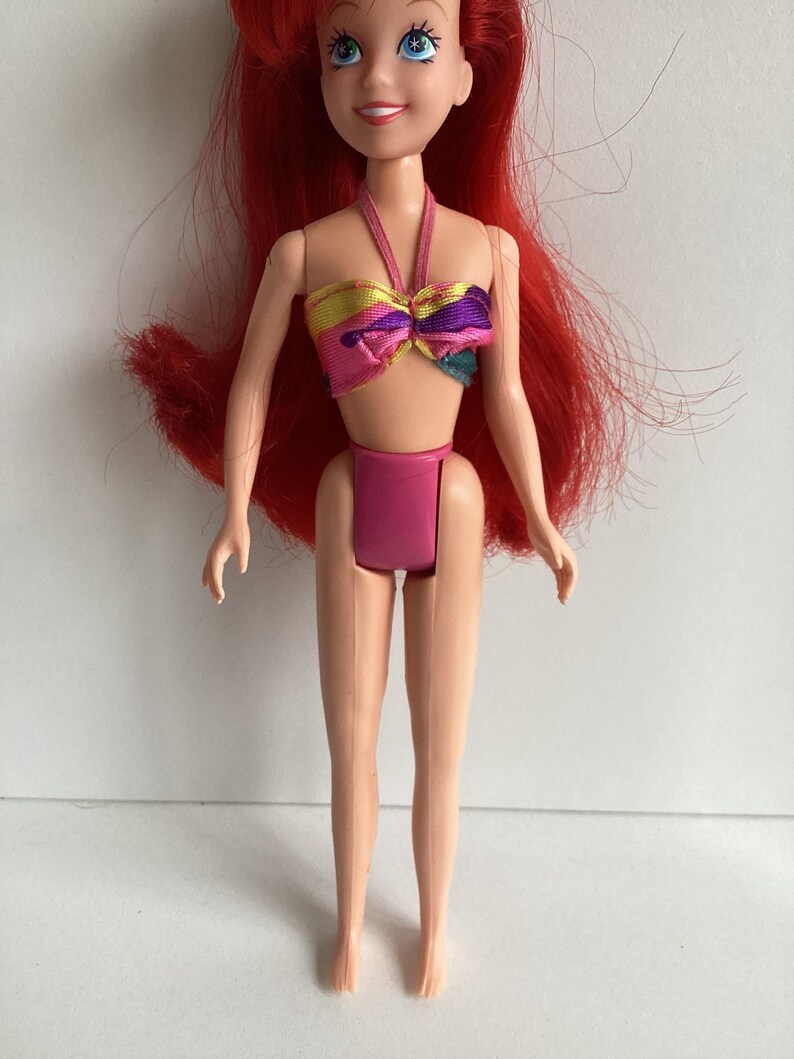 Peut inclure: Poup&eacute;e aux cheveux roux et aux yeux bleus, v&ecirc;tue d'un maillot de bain rose. Le haut de la poup&eacute;e est un style licou color&eacute; avec du jaune, du rose, du violet et du sarcelle. La poup&eacute;e est debout, les bras le long du corps.
