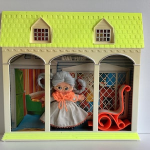 May include: A vintage dollhouse toy with a neon green roof and a white frame. Inside, a doll with gray hair, glasses, and an orange scarf stands in front of a window. The dollhouse is labeled "NANA FLATSY".