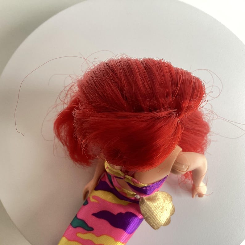 Peut inclure: Une poup&eacute;e avec des cheveux rouge vif et une robe color&eacute;e. La robe pr&eacute;sente un motif rose, jaune, violet et vert. Un accessoire dor&eacute; est attach&eacute; &agrave; la robe.