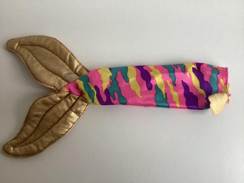 Peut inclure: Une queue de sir&egrave;ne jouet avec une nageoire dor&eacute;e et un corps &agrave; motif camouflage rose, jaune, turquoise et violet. La queue est con&ccedil;ue pour le jeu des enfants et les d&eacute;guisements imaginatifs.
