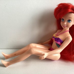 Peut inclure: Poup&eacute;e d'un personnage aux longs cheveux roux, aux yeux bleus et &agrave; l'expression souriante. La poup&eacute;e porte un haut et un bas de bikini color&eacute;s. La poup&eacute;e est assise.