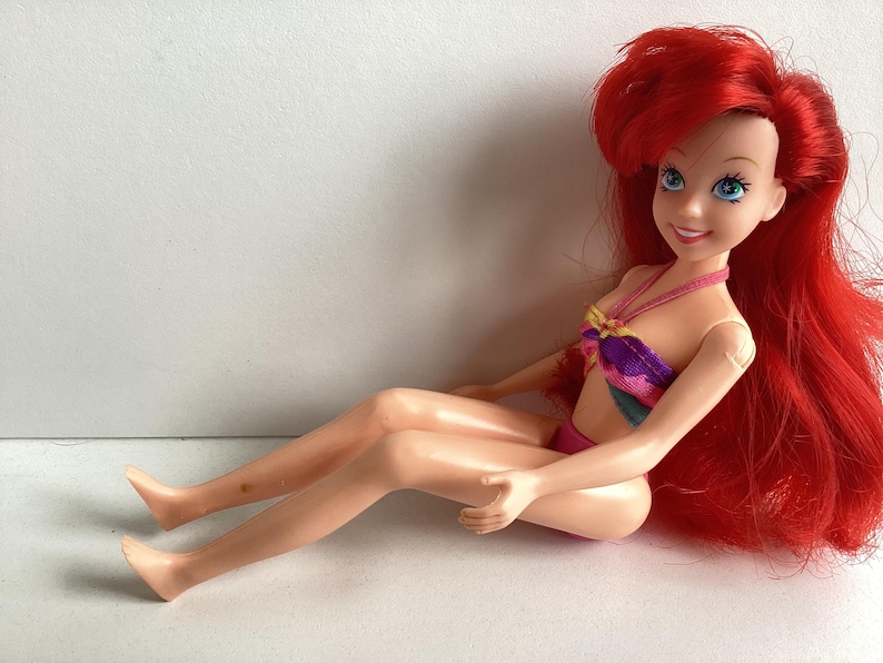 Peut inclure: Poup&eacute;e d'un personnage aux longs cheveux roux, aux yeux bleus et &agrave; l'expression souriante. La poup&eacute;e porte un haut et un bas de bikini color&eacute;s. La poup&eacute;e est assise.