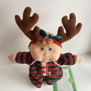May include: A Cabbage Patch Kids doll wearing a red and green striped onesie and a reindeer antler headband. The doll has blue eyes and a green bow. An adoption certificate is visible in the background.