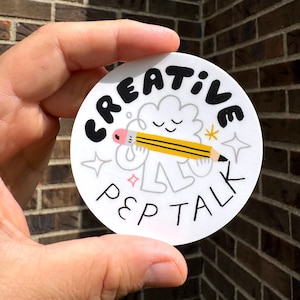 May include: A circular white sticker with the text "CREATIVE PEP TALK" in black. The design includes a smiling pencil and a cloud. The background is a brick wall.