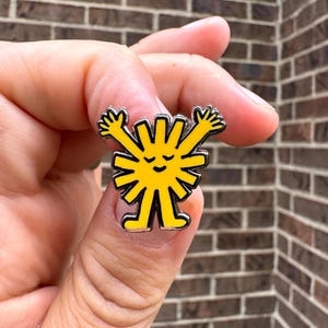 May include: A yellow enamel pin in the shape of a smiling sun with outstretched arms. The pin has a black outline and silver-colored trim. The pin is held in a hand against a brick background.