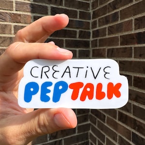 May include: A white sticker with the words "CREATIVE PEP TALK" in black, blue, and orange. The sticker is held in front of a brick wall.
