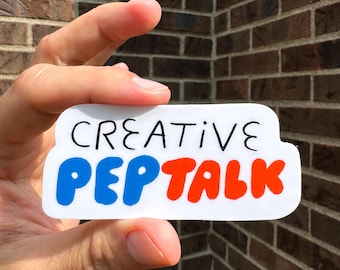 May include: A white sticker with the words "CREATIVE PEP TALK" in black, blue, and orange. The sticker is held in front of a brick wall.