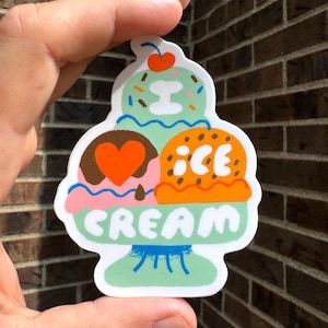 May include: A colourful sticker featuring a whimsical ice cream sundae design. The sticker includes three scoops of ice cream in different flavours, a cherry on top, and the word "CREAM" in green. The sticker is held against a brick wall.