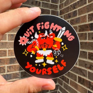 May include: Round black sticker featuring a cartoon demon illustration. The red demon holds a sword and flail, with the text "Quit Fighting Yourself" in pink and yellow. The sticker has a white border.