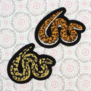 May include: Two embroidered snake patches. One features a brown and orange pattern with a white outline on a black background. The other patch has a yellow and black pattern with a black outline. Both patches are on a white background with a floral pattern.