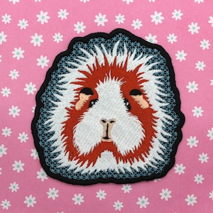 May include: Embroidered patch of a white and red guinea pig with a black outline. The guinea pig has a fluffy white mane and a red face.