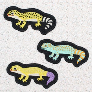 May include: Three embroidered gecko patches in yellow, blue, and yellow with purple tails. Each gecko is outlined in black and has a detailed design. The patches are on a speckled white background.