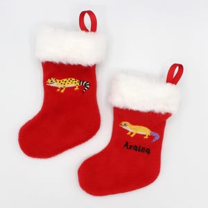 May include: Two red Christmas stockings with white faux fur trim and red hanging loops. Each stocking features an embroidered gecko design in yellow, black, and purple. One stocking is personalized with the name "Azalea".
