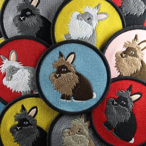 May include: A collection of nine embroidered patches featuring various fluffy bunnies in different colours and poses. The patches are circular and have a variety of backgrounds, including red, yellow, blue, and grey.
