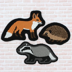 Puede incluir: Tres parches bordados con animales del bosque. El parche superior es un zorro naranja con pecho y cola blancos. El parche del medio es un erizo marrón. El parche inferior es un tejón gris y negro. Todos los parches tienen un borde negro.