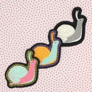 May include: Three embroidered snail patches in pink, orange, and grey with black borders. The snails have detailed stitching and are set against a pink background with a cross pattern. These patches are ideal for adding to clothing or accessories.
