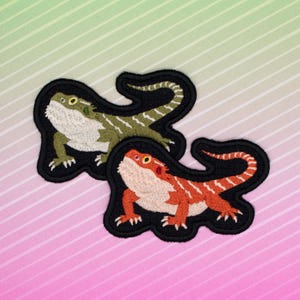 Peut inclure: Deux écussons brodés représentant des dragons barbus. L'un est vert et blanc, l'autre orange et blanc. Les deux ont une bordure noire et sont sur un fond rayé.