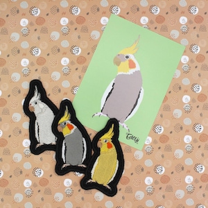 Puede incluir: Tres parches bordados para planchar de cacatúas en diferentes variaciones de color: blanco, gris y amarillo. Los parches están sobre un fondo marrón con lunares blancos y marrones. Una tarjeta con un dibujo de una cacatúa está en el fondo.