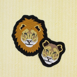Puede incluir: Dos parches bordados de león, uno más grande que el otro. El parche más grande es marrón con un contorno negro y presenta la cara de un león con melena. El parche más pequeño es marrón con un contorno negro y presenta la cara de un león con melena.