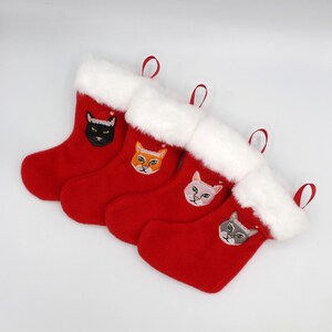 Op de afbeelding: Vier rode kerstsokken met witte bontkraag en geborduurde kattengezichten. De katten dragen kerstmanmutsen.