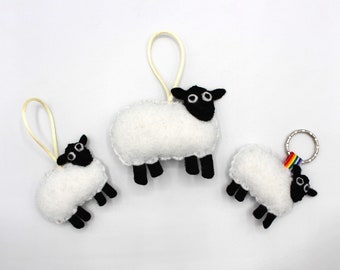 May include: Three felt sheep ornaments, one with a rainbow keyring. The sheep are white with black faces and eyes. The largest sheep has a loop for hanging.