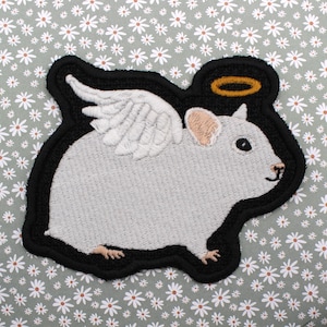 May include: A white embroidered patch of a hamster with wings and a halo. The hamster is smiling and has a black outline. The patch is on a green background with white daisies.