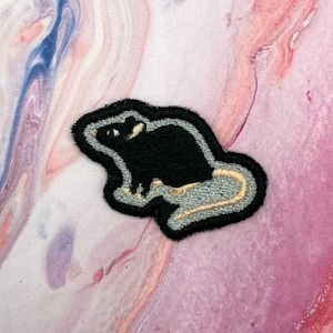 Peut inclure: Écusson brodé représentant un rat noir avec une queue grise et beige et un contour noir. L'écusson est posé sur un fond marbré rose et blanc. Idéal pour personnaliser vestes, sacs ou autres articles en tissu.