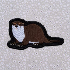 May include: An embroidered patch featuring a brown otter with a white chest and face, depicted in a side profile view. The otter has dark brown fur, a black nose, and small white claws, all outlined with a thick black border on a dark background.