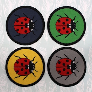 Peut inclure: Quatre patchs brodés thermocollants représentant une coccinelle rouge avec des points noirs et des accents blancs sur des fonds de couleurs différentes : bleu marine, vert, jaune et gris.