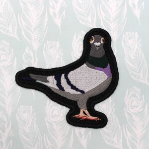 Puede incluir: Un parche bordado en blanco y negro de una paloma con cuello morado y verde. La paloma tiene ojos amarillos y un contorno negro.