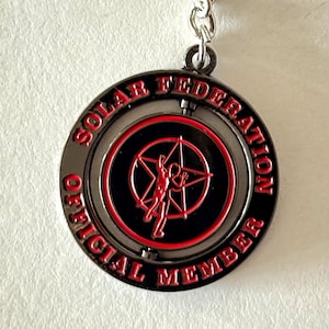 Puede incluir: Un llavero redondo con un anillo y una cadena plateados. El colgante es negro con un círculo interior rojo y una figura roja en forma de estrella. El texto "SOLAR FEDERATION OFFICIAL MEMBER" está en rojo alrededor del borde.