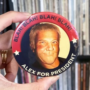 May include: A round button with a portrait of a man, the text "ALEX FOR PRESIDENT" in white, and the word "BLAH!" repeated around the edge. The button has a red and blue color scheme with white stars.